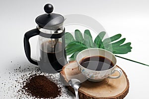French press coffee with cup of coffee spoon and coarse coffee powder on wooden pedestal with green leaf white background