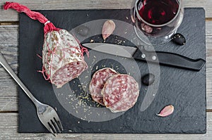 A french saucisson - dry summer sausage on the black background, with a knife, fork, garlic and olives and red wine.