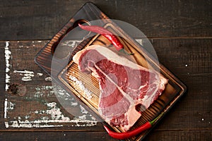 Fresh, aged t-bone steak on a cutting board with two pods of red chili peppers on a dark wooden background, place for text.