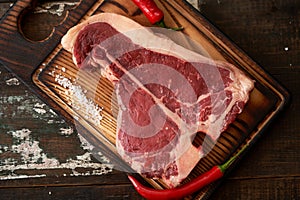 Fresh, aged t-bone steak on a cutting board with two pods of red chili peppers on a dark wooden background, place for text.