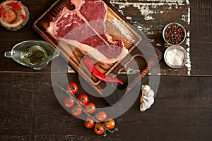 Fresh, aged t-bone steak on a cutting board with two red chili peppers, olive oil, dried tomatoes and paprika, salt and peppercorn