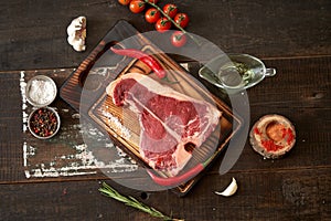 Fresh, aged t-bone steak on a cutting board with two red chili peppers, olive oil, dried tomatoes and paprika, salt and peppercorn