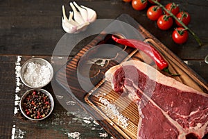 Fresh, aged t-bone steak on a cutting board with two red chili peppers, olive oil, dried tomatoes and paprika, salt and peppercorn