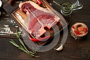 Fresh, aged t-bone steak on a cutting board with two red chili peppers, olive oil, dried tomatoes and paprika on a wooden dark