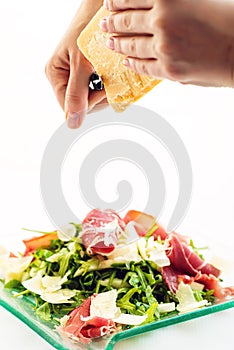 Fresh arugula vegetable salad with ham and cheese on glass plate on white background and cooks hands shredding cheese, pr