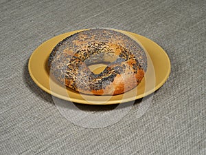 A fresh bagel with poppy seeds on a yellow plate on a rough gray tablecloth