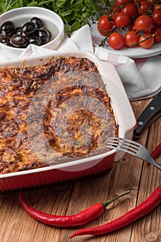 Fresh Baked Lasagne in Red Dish with Black Olives Tomatoes and Chilli on Wooden Table