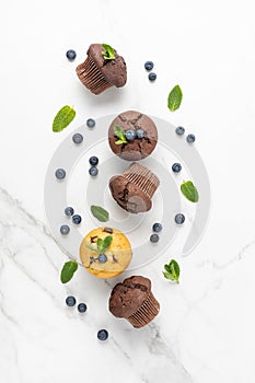Fresh baked muffins with chocolate chips, blueberry berries and mint leaves on white marble table background. Chocolate and banana