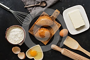 Fresh baked pasties and baking ingredients on a dark stone background. Russian pirozhki. Top view