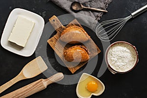 Fresh baked pasties and baking ingredients on a dark stone background. Russian pirozhki. Top view