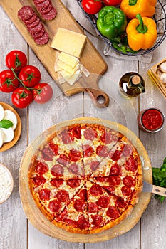 Fresh baked pepperoni pizza on wooden board and table, with colorful ingredients, vertical overhead top view