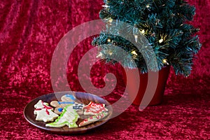 Fresh baked sugar cookies decorated by children for Christmas treats, on a wooden plate, with lighted tree, on a red background