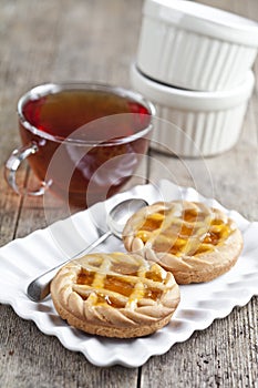 Fresh baked tarts with marmalade or apricot jam filling on white ceramic plate and cup of tea on rustic wooden table photo