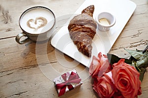 Fresh bakery croissant, coffee with heart sign, rose flowers on wooden table. Romantic breakfast for Valentine`s Day celebrate con