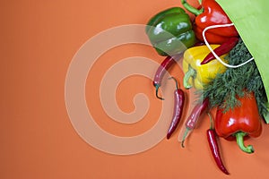 Fresh bell peppers in red, yellow and green colors, chili peppers and dill greens in a paper bag on an orange background. Bag for