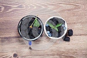 Fresh berries of blackberries, blueberries, mint leaves, in white bowl and blue mug on wooden background. Top view