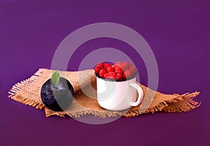 Fresh berries in a white vintage mug on burlap texture. Creative purple background with raspberries and plums. Close-up