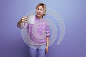 fresh blond young millennial woman in casual look holding out a cup of caffeinated drink on bright background with copy