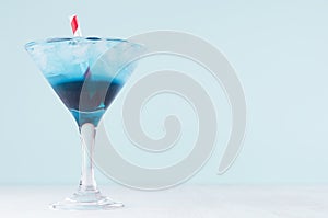 Fresh blue alcohol cocktail with ice cubes, red straw in misted wineglass on green color background, white wood board.