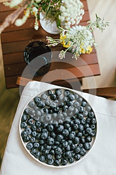 Fresh blueberries on modern ceramic plate, wildflowers bouquet in rustic room. Summer in countryside