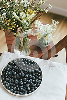 Fresh blueberries on modern ceramic plate and wildflowers bouquet in rustic room. Summertime in countryside. Healthy food