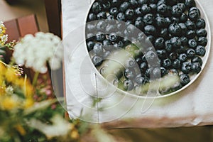 Fresh blueberries on modern ceramic plate and wildflowers bouquet in rustic room, top view.Aesthetic