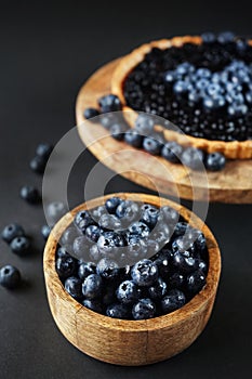 Fresh blueberries in a wooden bowl next to blueberry tart on a wooden stand