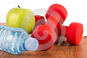 Fresh bottle of water with dew, green apple and red dumbbell on wooden floor