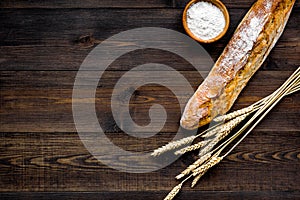 Fresh bread concept. Crispy french baguette near ears of wheat and bowl with flour on dark wooden background top view