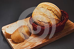 Fresh bread made from wheat flour in a wicker basket made of dark wood lies on a wooden board and against a dark background. Light