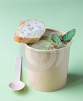 Fresh broccoli cream soup served with fresh bread, mint and spices in craft container on green background. Soup to go, healthy