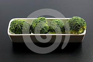 Fresh broccoli is housed in white ceramic vessels and placed on a black background closeup.