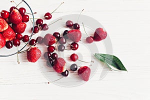 fresh cherries and strawberries scattered from bowl on white rustic wooden background. ripe juicy red berries on table, harvest