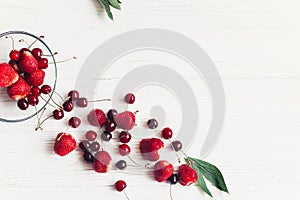 fresh cherries and strawberries scattered from bowl on white rustic wooden background. ripe juicy red berries on table, harvest