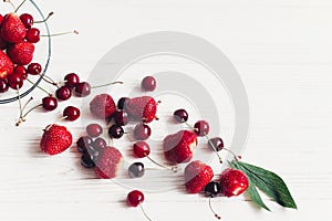 fresh cherries and strawberries scattered from bowl on white rustic wooden background. ripe juicy red berries on table, harvest