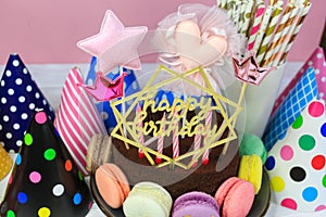 Fresh chocolate delicious birthday cake with macaroons and hats around it on table against pink background. Happy birthday.