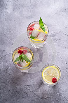 Fresh cold ice water drink with lemon, raspberry fruits and mint leaf in three glass on stone concrete background