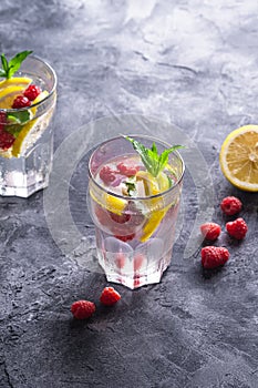 Fresh cold ice water drink with lemon, raspberry fruits and mint leaf in two faceted glass on stone concrete background