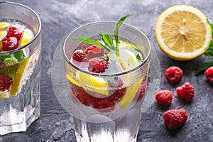 Fresh cold ice water drink with lemon, raspberry fruits and mint leaf in two faceted glass on stone concrete background