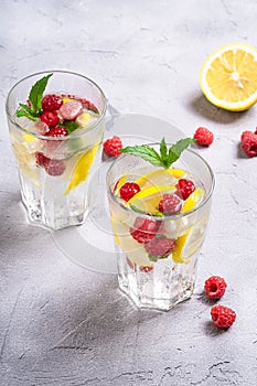 Fresh cold ice water drink with lemon, raspberry fruits and mint leaf in two faceted glass on stone concrete background