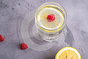 Fresh cold sparkling water drink with lemon, raspberry fruits in glass on stone concrete background, summer diet beverage