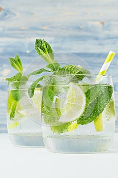 Fresh cold summer beverage gin and tonic with lime, leaf mint, straw, ice cubes, soda on light blue wooden background, closeup.