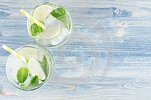 Fresh cold summer beverage mojito with lime, leaf mint, straw, ice cubes, soda on blue wood background, top view, border.