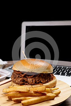 Fresh cooked beef burger with french fries in front of laptop against black background. Food delivery at home.