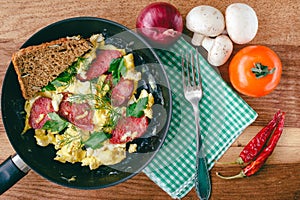 Fresh cooked scrambled eggs in pan with sausage and herbs. Bread, fork, vegetables, napkin on wooden board, top view.