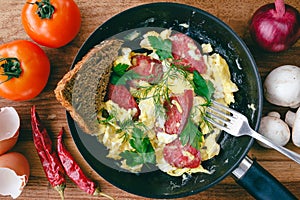 Fresh cooked scrambled eggs in pan with sausage and herbs. Bread, fork, vegetables on wooden board, top view.