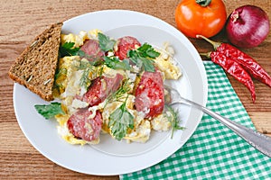 Fresh cooked scrambled eggs with sausage and herbs in white plate.Bread, napkin, fork, vegetables on wooden board.