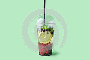 Fresh cool detox drink with Various berries in plastic cup on green background. Tasty infused water or lemonade to go. Proper