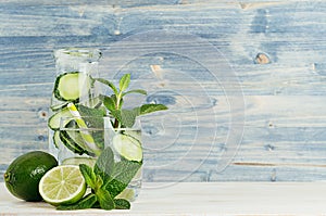 Fresh cool lemonade in bottle and glass with bubbles carbonated water, lime, cucumber, mint, ice on light blue wood plank.