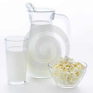 Fresh cow milk in a glass jug and cup in a next to a plate of fresh cottage cheese on white background.